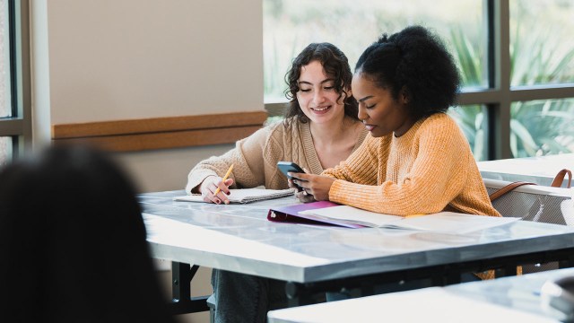 Two teenage girls take a break from their schoolwork to play with a smart phone.